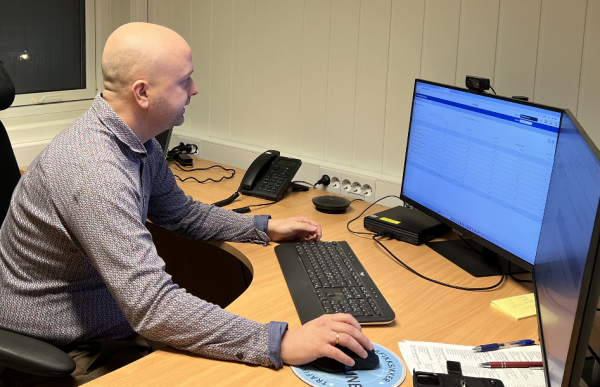 En mann i en lys mønstret skjorte sitter på et kontor foran en PC med to skjermer. Foran han ligger det noen papirer, to penner og en mobiltelefon.