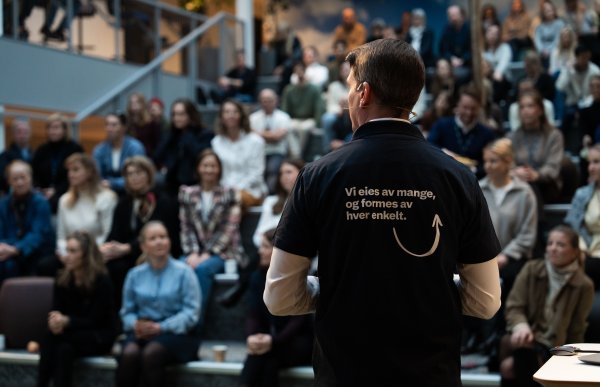 Mann med mørk t-skjorte over en hvit genser står på en scene og holder et foredrag foran mange mennesker. Bildet er tatt bakfra, han har en mygg-mikrofon for å snakke. På ryggen står teksten "Vi eies av mange, men formes av hver enkelt" på t-skjorta.