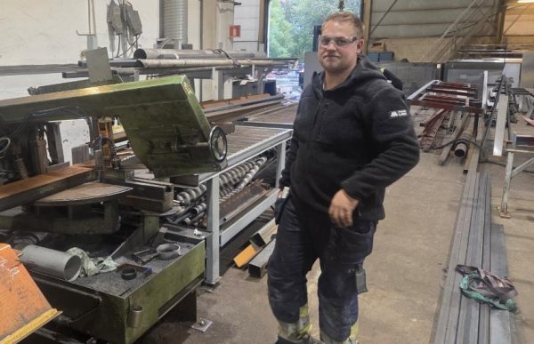 En mann står inne i et verksted omgitt av metallplater, rør og store maskiner. Han har på seg vernebriller, svart arbeidsjakke og mørke arbeidsbukser med refleksdetaljer. I bakgrunnen er det åpne porter som slipper inn dagslys og viser trær utenfor. Verkstedet er fullt av industrielt utstyr, og gulvet er støvete etter arbeid.