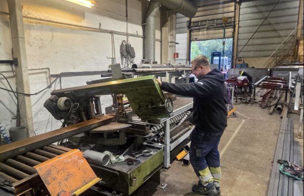 En mann står inne i et verksted og jobber ved en stor metallmaskin. Han har på seg arbeidsklær med mørk genser, arbeidsbukse med gule refleksfelt og vernesko. Han holder i en grønn maskindel, og det ligger en lang metallstang på rullebord foran maskinen. I bakgrunnen er det flere metallrør. Rommet er stort med høyt tak, industriell belysning og en åpen port ut mot dagslys.