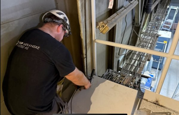 En mann jobber inne i et industrielt anlegg. Han har på seg vernehjelm, vernebriller og en svart T-skjorte med firmalogo på ryggen. Han bøyer seg frem og arbeider med verktøy på en metallflate nær et kabelsystem som går langs veggen. Rundt ham er det stålkonstruksjoner, rør og teknisk utstyr, og området er opplyst av arbeidsbelysning.