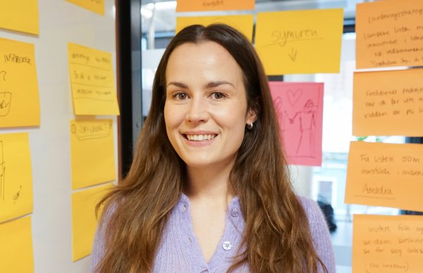 Kvinne med langt, brunt hår i lilla cardigan. Hun ser rett i kameraet og smiler. I bakgrunnen er en tavle og en glassdør dekket med gule og noen rosa post-it-lapper.