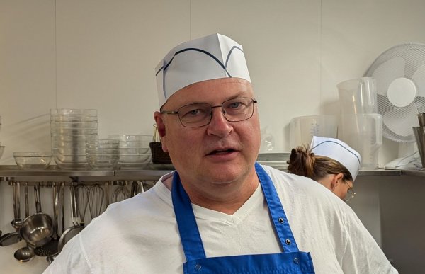 En mann med briller, hvit t-skjorte, blått forkle og hvit hatt. I bakgrunnen skimtes kjøkkenredskaper. 