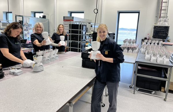 Fire damer står rundt et bord i et stort rom eller et slags verksted. De har noen hvite, små roboter på bordet foran seg. Noen av dem holder de hvite robotene opp for å vise de fram.