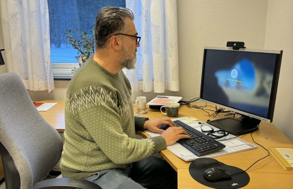 Mann med grønn genser og briller sitter foran en pc på et kontor. Han har en hjørnepult.
