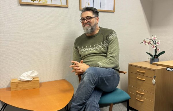 Mann med jeans og grønn genser og briller sitter på en stol med et lite, lavt bord ved siden av seg. Det står en pakke lommetørklær på bordet og henger noen illustrasjoner på veggene. 