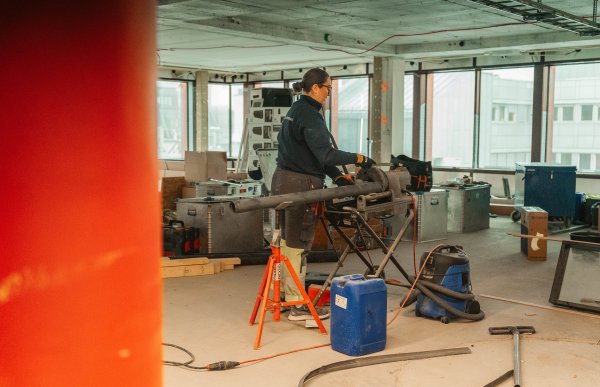 En kvinne står inne på en byggeplass med en mellomstor maskin. Hun arbeider med et rør, og holder på å frese inn et spor i enden.