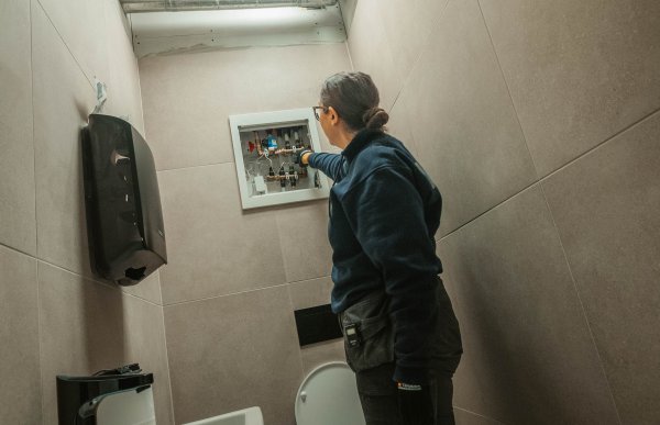 En kvinne i arbeidsklær står inne på et toalett. Hun har hånden inne i et rør-i-rør-skap og ser ut som hun tester at det fungerer.