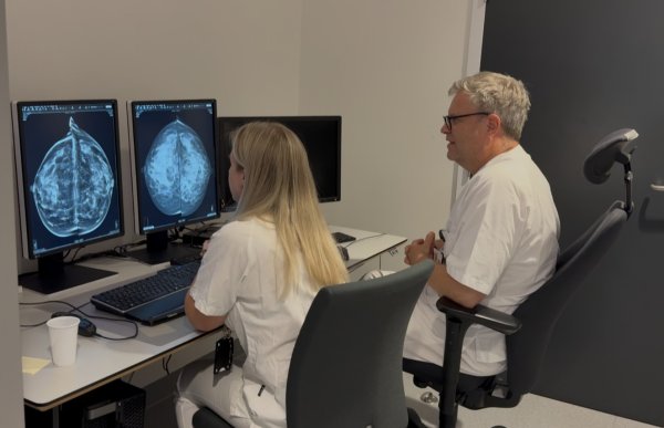 En kvinne og en mann i hvitt arbeidstøy sitter sammen foran to skjermer med mammografibilder. De ser på bildene og snakker sammen. Rommet er innredet med arbeidsstasjon, tastatur og flere dataskjermer.