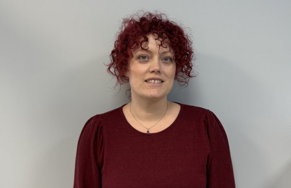 En kvinne med kort, krøllete rødt hår står foran en lys, nøytral vegg og ser rett mot kamera. Hun har på seg en mørk rød genser med lange ermer og et enkelt smykke rundt halsen. Ansiktsuttrykket er rolig og vennlig med et svakt smil.