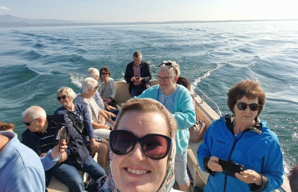 Dame med solbriller og skjerf på hodet sitter i en båt og bak i båten sitter en gjeng pensjonister. Det er sol og fint vær på sjøen. 