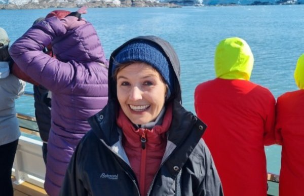 En kvinne smiler mot kameraet på en båt. Hun har på seg vinterklær med lue og hette, og i bakgrunnen ser man flere personer i fargerike jakker som ser utover vannet mot en stor isbre.