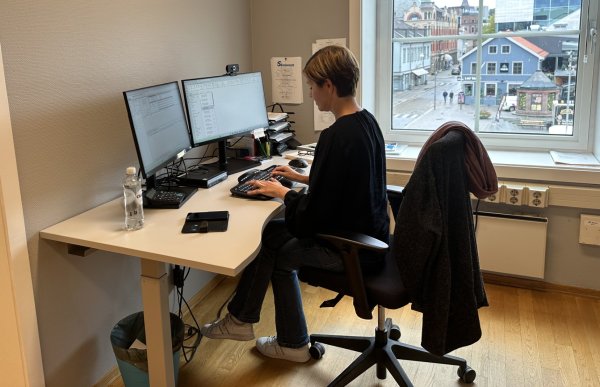 En kvinne sitter ved et skrivebord på kontor og jobber på to dataskjermer. Hun har mørk overdel og lyse jeans, og på stolen henger en jakke. Utenfor vinduet ser man en bygate med bygninger og mennesker.