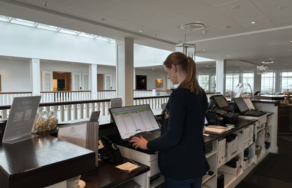 En kvinne står bak en lang skranke. Hun har brun hestehale og har på seg en mørkeblå dressjakke. Hun står foran en PC som viser en slags tabell. Foran henne kan man se et hvitt rekkverk. Ved siden av henne er flere andre skjermer og en hylle med diverse permer og annet utstyr.