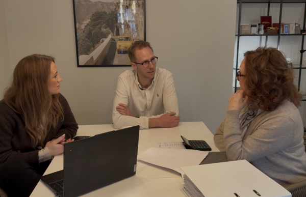 Tre personer sitter rundt et bord i et kontor og snakker sammen. En mann med briller sitter mellom to kvinner og lener seg litt frem mens han snakker. På bordet ligger en laptop, en kalkulator, en perm og dokumenter. På veggen bak henger et bilde av en vei med en gul bil.