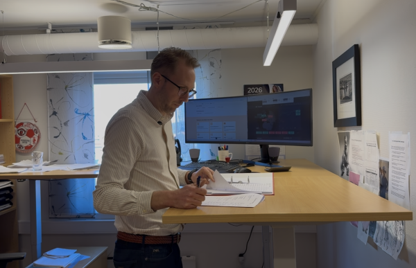 En mann med en lys, stripete skjorte står ved et hev- og senkbart skrivebord og ser ned på et ark han holder i hånden. Han skriver eller markerer på papiret med en penn. Bak ham står en stor dataskjerm, og rundt skrivebordet ligger det papirer og kontorutstyr.