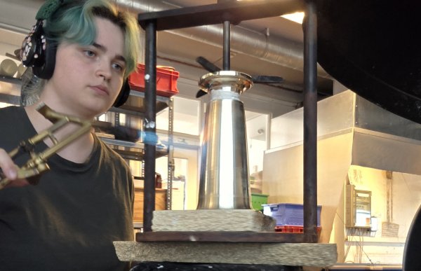 En kvinne med headset og grønt hår står, ved en arbeidsbenk på et verksted. Hun fører flammer mot en sølvgjenstand. Rundt henne er det arbeidsbenker, metallstenger og teknisk utstyr. 