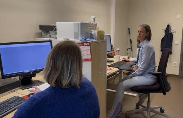 To kvinner sitter ved datamaskiner på et kontor. Kvinnen i forgrunnen har kort, lyst hår og en blå genser. Kvinnen i bakgrunnen har halvlangt, brunt hår, lys blå skjorte og lyse bukser. Hun vender seg mot kollegaen mens de ser ut til å snakke sammen.