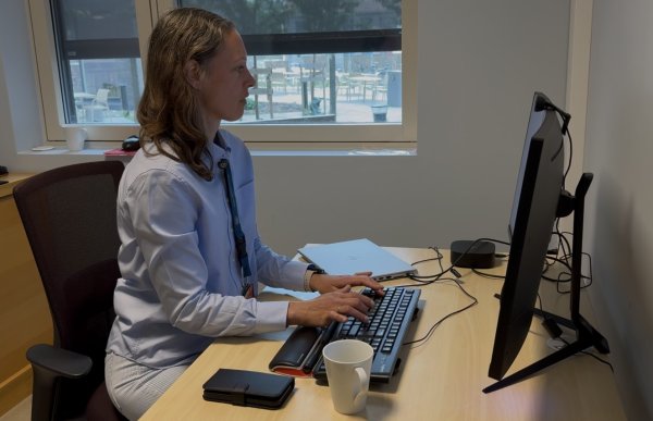 En kvinne med halvlangt, brunt hår sitter ved en kontorpult og arbeider på en datamaskin. Hun har på seg en lys blå skjorte og lyse bukser. På skrivebordet står blant annet en kaffekopp, en mobiltelefon og en lukket PC.