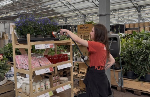En ung kvinne med langt, mørkebrunt hår vanner blomster i et hagesenter med en vannslange. Hun har på seg en rød T-skjorte og en svart genser knyttet rundt livet og konsentrerer seg om å vanne plantene på utstillingen.