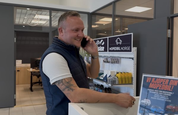 En mann står ved en disk i et moderne kontor- og salgslokale og snakker i mobiltelefon. Han har på seg en hvit T-skjorte og mørkeblå vest. Bak ham er det kontorer med glassvegger og en hylle med produkter og skilt fra Mopedbil Norge.