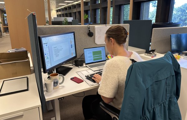 En kvinne med lys genser og oppsatt hår sitter ved en kontorplass foran to skjermer, en stor og en bærbar PC. Hun skriver på tastaturet og jobber med en presentasjon. På pulten står flere kaffekopper, en mobiltelefon og en notatblokk. Hun har en mørkeblå jakke hengende over stolryggen.