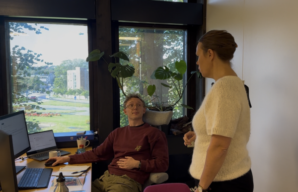 En kvinne med lys genser og oppsatt hår står ved en mann som sitter foran en dataskjerm ved et skrivebord. Mannen har briller og rødlilla genser. Han ser opp på henne mens hun snakker. Bak dem er det et stort vindu med grønne planter på vinduskarmen og utsikt over en vei og bygninger utenfor.
