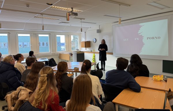 En kvinne står foran en klasse og holder et foredrag i et klasserom. Elevene sitter ved pulter og ser mot henne og et stort lerret. På lerretet vises en presentasjon med teksten «SEX VS PORNO». Det er dagslys fra store vinduer, og rommet har typisk skoleinventar.
