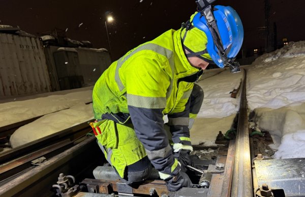 En mann med gule arbeidsklær med refleks, blå vernehjelm og lykt justerer sporvekslingen i togsporet. Det er snø og vinter rundt. Mørkt. 