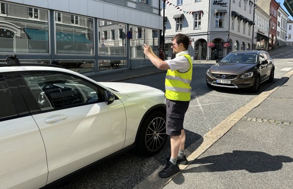 En mann i gul refleksvest over en hvit t-skjorte, svarte shorts, svarte sko og grå sokker holder henda opp fordi han tar et bilde av en bil med telefonen sin. Han har på seg solbriller, det er blå himmel og bygårder i bakgrunn. 