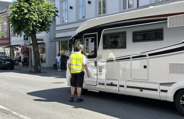 En mann står med ryggen til og prater med en annen som sitter inni en bobil. Vinduet til bobilsjåføren er nede, så de prater gjennom vinduet. Mannen som står har på seg en gul refleksvest hvor det står "parkering" på ryggen, svarte shorts og svarte sko. I bakgrunn er det en bygård som er hvit og grå, blå himmel og et grønt tre. Bobilen er hvit og svart. 