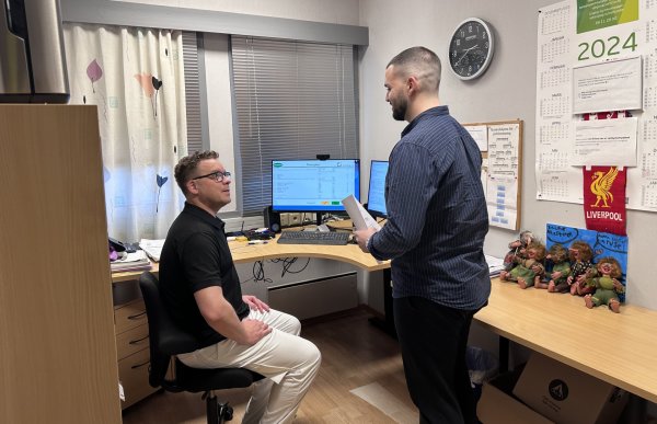 To menn er på et kontor. Den ene, med mørkt hår og skjegg, står og holder et ark mens han snakker med en kollega som sitter foran en dataskjerm. Den sittende mannen har kort, krøllete hår og briller, og han er kledd i sort skjorte og lyse bukser. På veggen bak dem henger kalendere, en Liverpool-vimpel og flere små trollfigurer.