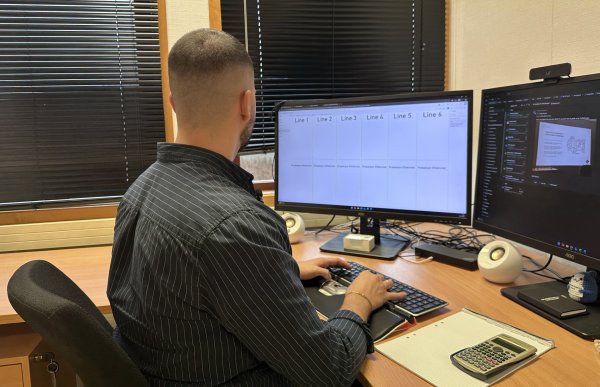 En mann med kort mørkt hår og skjegg sitter ved et skrivebord og jobber på en datamaskin med to store skjermer. Han har på seg en mørkeblå skjorte med hvite striper. På skrivebordet ligger en kalkulator, et notatark og ulike kontorrekvisita. Rommet har dempet belysning og mørke persienner foran vinduet.