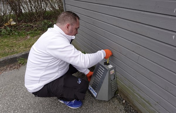 Mann med hvit genser sitter på huk foran en rottekasse som er montert fast på en vegg.