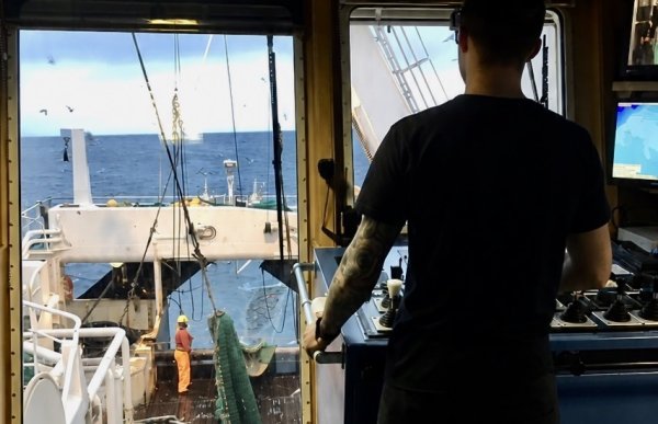 En mann i svarte klær står med ryggen mot kameraet foran et kontrollpanel. Gjennom store vinduer sees dekket, en person i oransje arbeidstøy, fiskeredskaper og åpent hav.