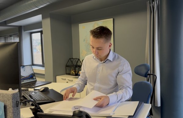 En mann med kort, lyst hår og lyseblå skjorte står ved et skrivebord på et kontor og blar i dokumenter i en perm. På skrivebordet er en dataskjerm, en bærbar PC og en telefon.