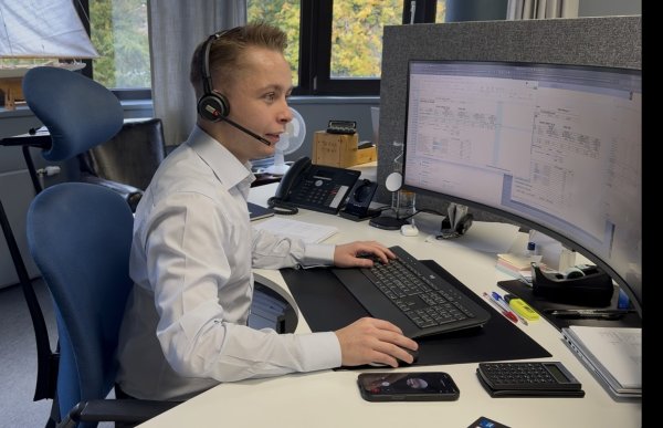 En mann med kort, lyst hår og lyseblå skjorte sitter i en blå kontorstol foran en stor, buet dataskjerm. Han har på seg hodetelefoner med mikrofon og arbeider ved tastaturet og datamusen. På skjermen vises flere dokumenter med tabeller og tall.