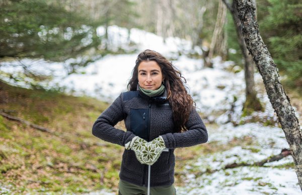 Dame med mørkt hår og svart fleecejakke og hjemmestrikka vottar står utendørs i et delvis snødekt skogsområde. Hun har et tynt redskap med begge hender foran seg.