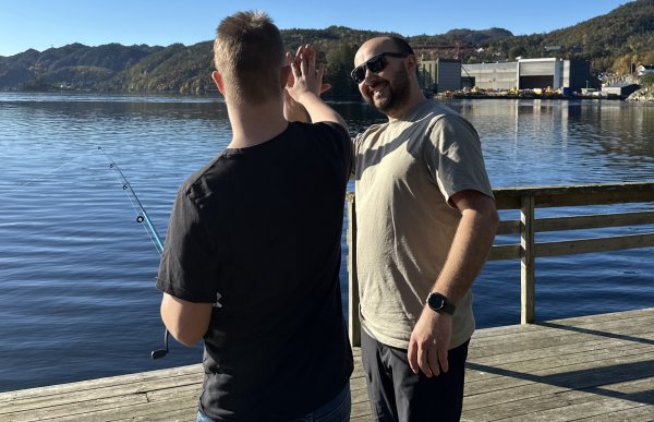Skoleassistent Tommy Egeland gir ros til en elev og smiler To personer står på en brygge og gir hverandre high-five. Det er sol og blå himmel. Den ene personen som står på med ryggen til, har en fiskestang i hånda.