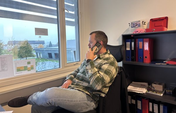 En mann sitter i et kontor ved et vindu og snakker i telefonen. Han har beina i kors og ser ut av vinduet. På veggen bak ham står en hylle med permer og kontorutstyr. Utenfor vinduet sees et skoleområde med bygninger og trær.