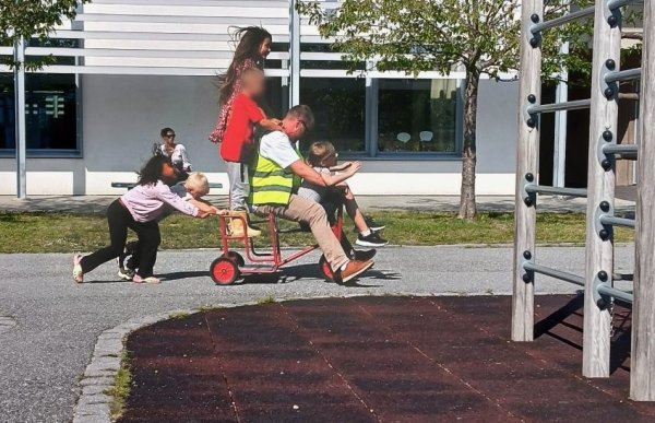 Bildet viser en mann med gul refleksvest som sitter på en trehjulssykkel sammen med et barn foran seg. To barn hjelper til med å skyve sykkelen bakfra, mens to barn står bak på rammen. De er utendørs på et lekeområde ved en skole med trær og et bygg i bakgrunnen. 