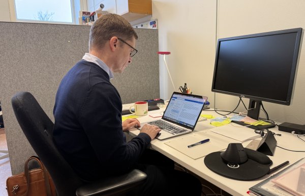 En mann sitter ved et skrivebord på et kontor og jobber på en bærbar datamaskin. Han har kort hår, briller og er kledd i en mørk genser over en lys skjorte. På pulten ligger blant annet en kaffekopp, notater, penn og en datamus. Ved siden av står en ekstern skjerm som er avslått. Bak ham er en skillevegg og et vindu som slipper inn dagslys.