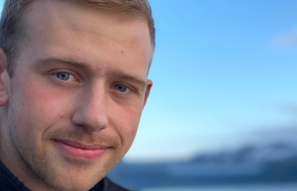 Bildet er et nært portrett av en ung mann som smiler svakt mot kameraet. Han har kort, lyst hår, blå øyne og lett skjeggvekst. Han har på seg en mørk jakke. Bak ham ser man havet og et blålig landskap i myk uskarp bakgrunn, trolig fjell i det fjerne. Himmelen er klar og blå.