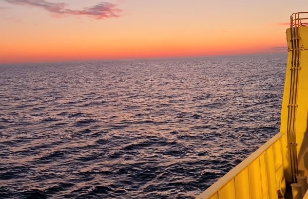 Du ser ut over et åpent hav fra dekket på en båt. Havet fyller mesteparten av bildet, med små bølger som beveger seg rolig og fanger opp lyset fra solnedgangen. Horisonten er rett og klar. Over den ligger en intens, varm himmel i sterke nyanser av oransje, rosa og mykt lilla. En liten skyformasjon øverst til venstre er svakt rosa i kanten. Til høyre i bildet vises en del av båtens side i gult metall, opplyst av det varme kveldslyset.