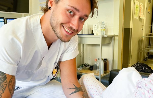 Sykepleier Erik Tørud er i ferd med å ta en blodprøve av en pasient på et sykehusrom. Vi ser bare armen til pasienten. Tørud smiler til kameraet. Han har mørkblondt hår, litt skjegg og bart. 