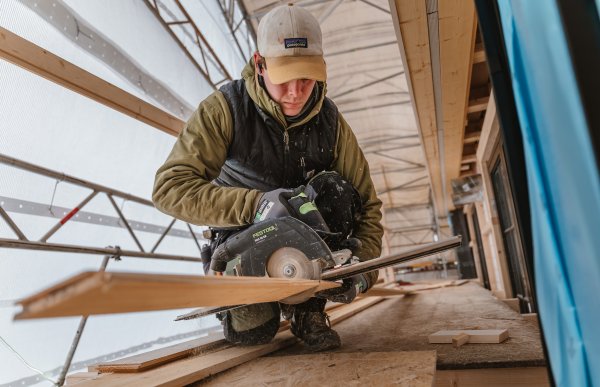 En mann sitter på knær inne på en byggeplass. I hendene har han en sirkelsag som han kapper en planke med. Han ser konsentrert ned, og capsen dekker til deler av ansiktet.