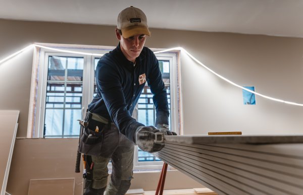 En mann i arbeidsklær og caps står innendørs på et byggeprosjekt og pusser kanten på en gipsplate med en gipshøvel. I bakgrunnen er et vindu til utsiden, og det henger en lyslenke på veggen.