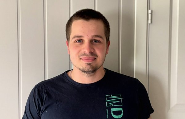 En mann med svart hår, skjegg og bart smiler. Han har på seg en svart t-skjorte med lys blå skrift. Bak mannen er det en grå panelvegg.