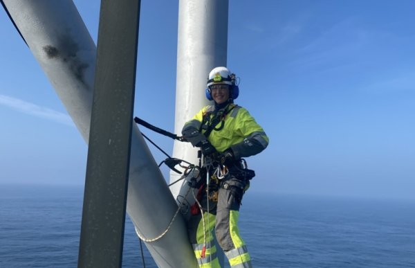 En dame i gule refleks arbeidsklær er spennet fast i sikringsutstyr til store sølvfargede stolper i høyden. Bak hun er det åpent hav, og stolpene er på en oljeplattform. Hunn har på seg hjelm, briller og hørselsvern, og smiler bredt.