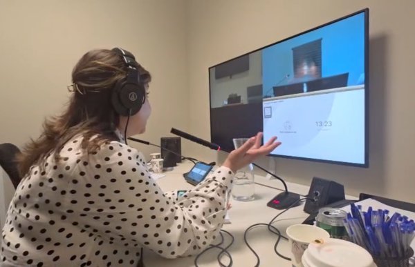 En kvinne med hodetelefoner og en hvit bluse med svarte prikker sitter ved et skrivebord foran en stor skjerm. På skjermen vises en person via video. Kvinnen snakker i en mikrofon og gestikulerer med den ene hånden.